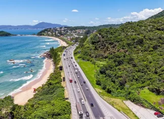Operação Verão: Autopista Litoral Sul prevê 5,4 milhões de veículos entre Natal e Ano Novo Imagem aérea mostra trecho da BR-101 à beira-mar, com fluxo de veículos em rodovia cercada por vegetação e praia, ilustrando o movimento intenso previsto na Operação Verão.