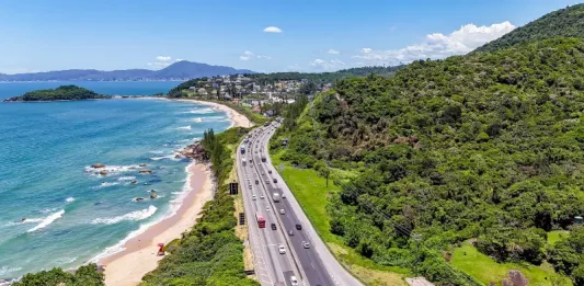 Imagem aérea mostra trecho da BR-101 à beira-mar, com fluxo de veículos em rodovia cercada por vegetação e praia, ilustrando o movimento intenso previsto na Operação Verão.