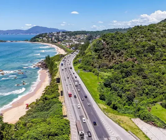 Operação Verão: Autopista Litoral Sul prevê 5,4 milhões de veículos entre Natal e Ano Novo Imagem aérea mostra trecho da BR-101 à beira-mar, com fluxo de veículos em rodovia cercada por vegetação e praia, ilustrando o movimento intenso previsto na Operação Verão.