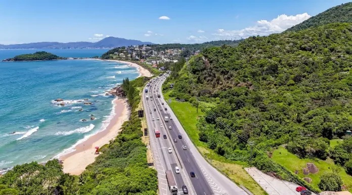Imagem aérea mostra trecho da BR-101 à beira-mar, com fluxo de veículos em rodovia cercada por vegetação e praia, ilustrando o movimento intenso previsto na Operação Verão.