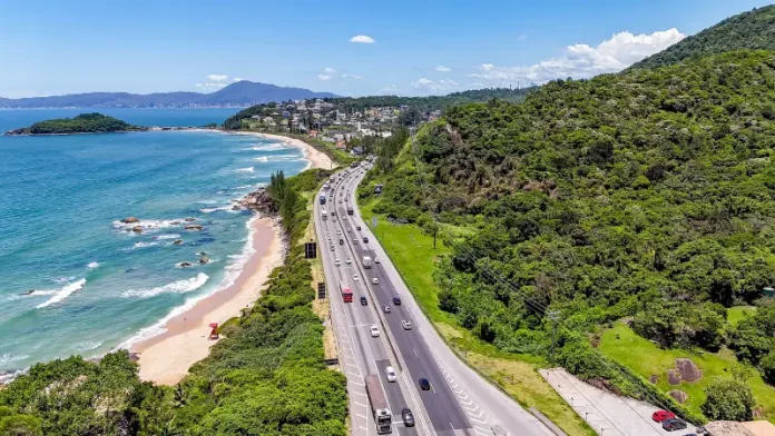 Operação Verão Imagem aérea mostra trecho da BR-101 à beira-mar, com fluxo de veículos em rodovia cercada por vegetação e praia, ilustrando o movimento intenso previsto na Operação Verão.
