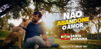 Homem sentado em um parque abraça seu cachorro; banner da campanha do Governo de SC sobre abandono de animais ao fundo, destacando a conscientização e o cuidado com os pets.