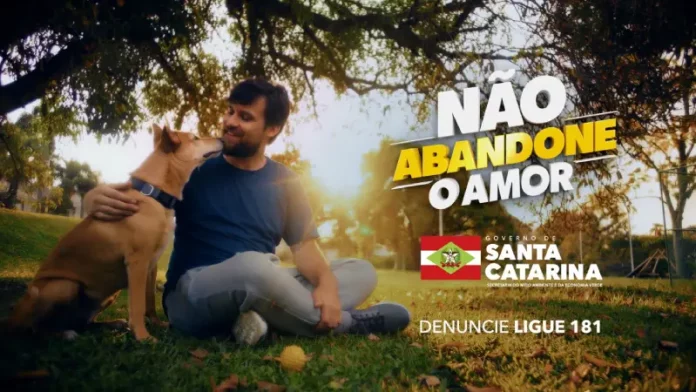 Homem sentado em um parque abraça seu cachorro; banner da campanha do Governo de SC sobre abandono de animais ao fundo, destacando a conscientização e o cuidado com os pets.
