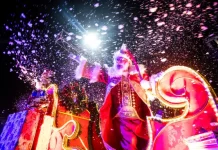 Imagem mostra Papai Noel em um carro alegórico iluminado durante o Desfile de Natal de Florianópolis, com efeitos de neve artificial e luzes, destacando o clima festivo do evento na Beira-Mar Norte.