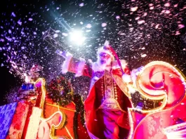 Imagem mostra Papai Noel em um carro alegórico iluminado durante o Desfile de Natal de Florianópolis, com efeitos de neve artificial e luzes, destacando o clima festivo do evento na Beira-Mar Norte.