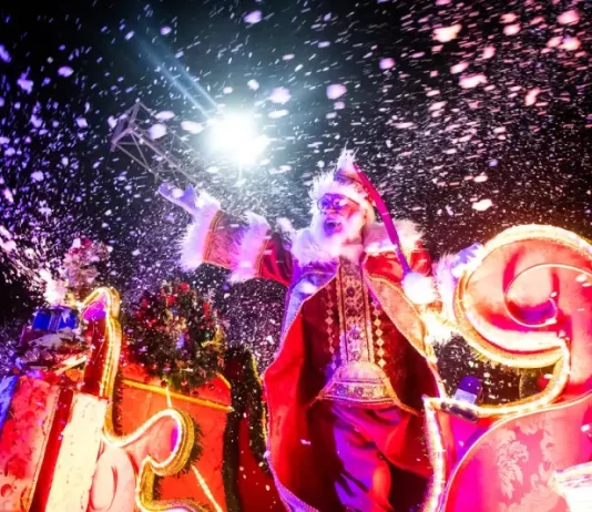 Imagem mostra Papai Noel em um carro alegórico iluminado durante o Desfile de Natal de Florianópolis, com efeitos de neve artificial e luzes, destacando o clima festivo do evento na Beira-Mar Norte.