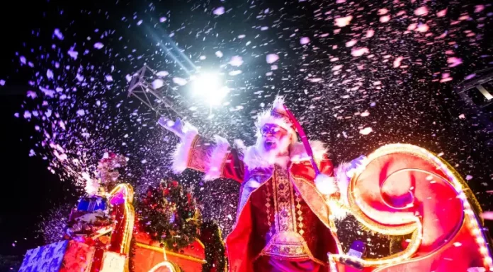 Imagem mostra Papai Noel em um carro alegórico iluminado durante o Desfile de Natal de Florianópolis, com efeitos de neve artificial e luzes, destacando o clima festivo do evento na Beira-Mar Norte.