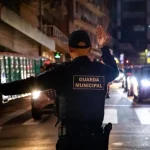 Agente da Guarda Municipal de Florianópolis orienta o trânsito à noite em via urbana, com veículos parados e iluminação pública ao fundo.