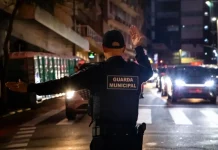 Agente da Guarda Municipal de Florianópolis orienta o trânsito à noite em via urbana, com veículos parados e iluminação pública ao fundo.