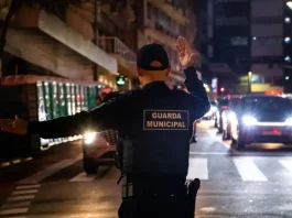 Agente da Guarda Municipal de Florianópolis orienta o trânsito à noite em via urbana, com veículos parados e iluminação pública ao fundo.