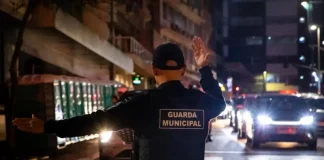 Agente da Guarda Municipal de Florianópolis orienta o trânsito à noite em via urbana, com veículos parados e iluminação pública ao fundo.