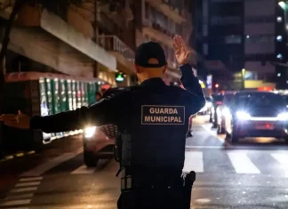 Agente da Guarda Municipal de Florianópolis orienta o trânsito à noite em via urbana, com veículos parados e iluminação pública ao fundo.