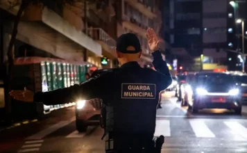 Agente da Guarda Municipal de Florianópolis orienta o trânsito à noite em via urbana, com veículos parados e iluminação pública ao fundo.