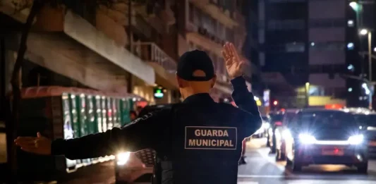 Agente da Guarda Municipal de Florianópolis orienta o trânsito à noite em via urbana, com veículos parados e iluminação pública ao fundo.
