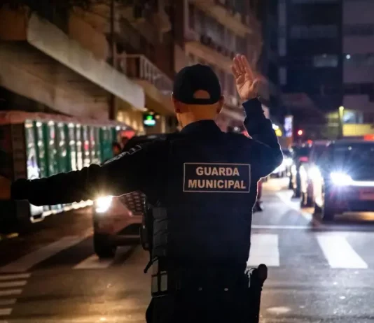 Agente da Guarda Municipal de Florianópolis orienta o trânsito à noite em via urbana, com veículos parados e iluminação pública ao fundo.