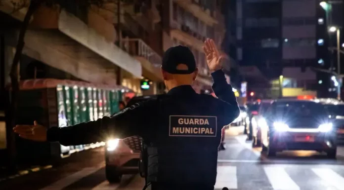 Agente da Guarda Municipal de Florianópolis orienta o trânsito à noite em via urbana, com veículos parados e iluminação pública ao fundo.
