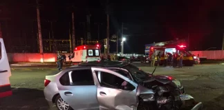 Carro com a frente destruída após colisão frontal na SC-405, enquanto equipes do Corpo de Bombeiros e do SAMU realizam atendimento às vítimas durante a noite.