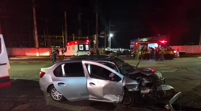 Carro com a frente destruída após colisão frontal na SC-405, enquanto equipes do Corpo de Bombeiros e do SAMU realizam atendimento às vítimas durante a noite.