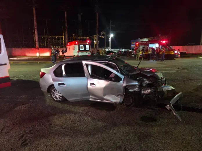 Carro com a frente destruída após colisão frontal na SC-405, enquanto equipes do Corpo de Bombeiros e do SAMU realizam atendimento às vítimas durante a noite.