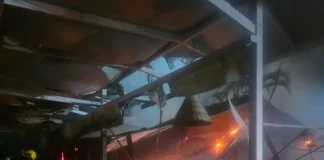 Foto mostra bombeiros atuando no combate ao incêndio dentro do restaurante instalado em uma marina, destacando as chamas ainda ativas e a destruição parcial da estrutura.