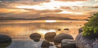 Imagem utilizada para ilustrar a mudança no tempo antes da formação do ciclone extratropical, mostrando um amanhecer calmo em uma praia, com pedras na água, céu parcialmente nublado e o sol refletindo no mar, simbolizando a transição entre estabilidade e o avanço das instabilidades