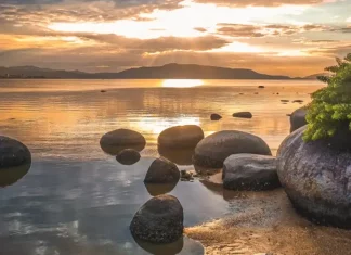 Domingo com sol e ciclone extratropical ao longo da semana Imagem utilizada para ilustrar a mudança no tempo antes da formação do ciclone extratropical, mostrando um amanhecer calmo em uma praia, com pedras na água, céu parcialmente nublado e o sol refletindo no mar, simbolizando a transição entre estabilidade e o avanço das instabilidades