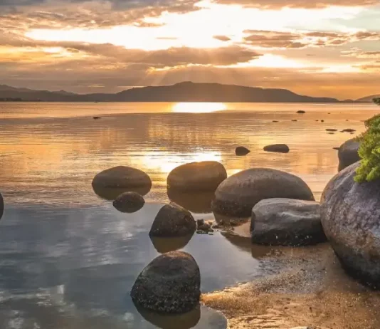 Domingo com sol e ciclone extratropical ao longo da semana Imagem utilizada para ilustrar a mudança no tempo antes da formação do ciclone extratropical, mostrando um amanhecer calmo em uma praia, com pedras na água, céu parcialmente nublado e o sol refletindo no mar, simbolizando a transição entre estabilidade e o avanço das instabilidades