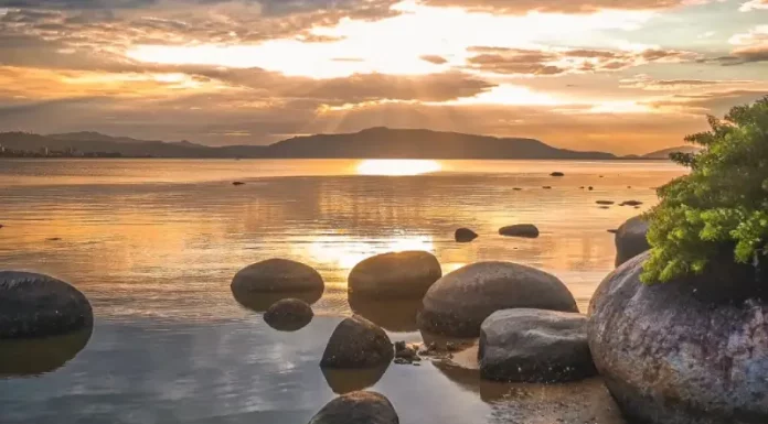 Imagem utilizada para ilustrar a mudança no tempo antes da formação do ciclone extratropical, mostrando um amanhecer calmo em uma praia, com pedras na água, céu parcialmente nublado e o sol refletindo no mar, simbolizando a transição entre estabilidade e o avanço das instabilidades