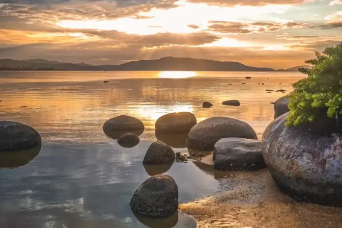 praia-da-Guararema-1 Imagem utilizada para ilustrar a mudança no tempo antes da formação do ciclone extratropical, mostrando um amanhecer calmo em uma praia, com pedras na água, céu parcialmente nublado e o sol refletindo no mar, simbolizando a transição entre estabilidade e o avanço das instabilidades