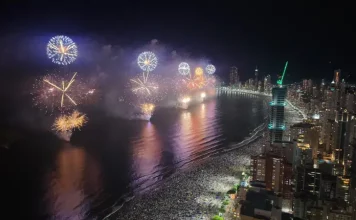 Imagem aérea do Réveillon em Balneário Camboriú, mostrando a queima de fogos sobre a Praia Central lotada e a orla iluminada durante a virada do ano.