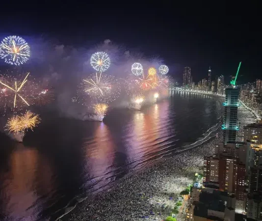 Imagem aérea do Réveillon em Balneário Camboriú, mostrando a queima de fogos sobre a Praia Central lotada e a orla iluminada durante a virada do ano.