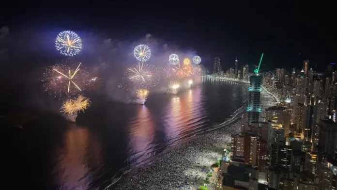 Imagem aérea do Réveillon em Balneário Camboriú, mostrando a queima de fogos sobre a Praia Central lotada e a orla iluminada durante a virada do ano.