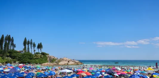 Semana começa com tempo firme e termina com calor intenso e temporais em Santa Catarina Imagem ilustra dia de calor intenso no litoral de Santa Catarina, com praia lotada, guarda-sóis abertos e grande concentração de pessoas aproveitando o tempo firme.