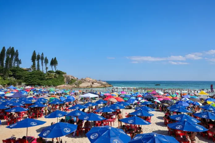 Imagem ilustra dia de calor intenso no litoral de Santa Catarina, com praia lotada, guarda-sóis abertos e grande concentração de pessoas aproveitando o tempo firme.