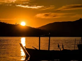 Semana terá tempo estável e amanheceres mais frios em SC Imagem ilustrativa de um pôr do sol sobre um corpo d’água, com barco em silhueta ao fundo, utilizada para representar condições de tempo estável, clima agradável e transição do dia para a noite.