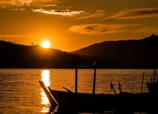 Imagem ilustrativa de um pôr do sol sobre um corpo d’água, com barco em silhueta ao fundo, utilizada para representar condições de tempo estável, clima agradável e transição do dia para a noite.