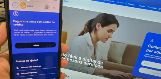 Celesc libera pagamento da conta de luz com cartão de crédito e parcelamento em até 21 vezes Aplicativo da Celesc exibido em um celular mostra a opção de pagamento da conta de luz com cartão de crédito, enquanto ao fundo aparece a Agência Web da companhia, ilustrando os canais digitais disponíveis para os clientes.