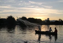 Barco pequeno com pescadores artesanais no Rio Biguaçu ao entardecer, com um deles lançando a rede sobre a água enquanto o sol se põe ao fundo, formando silhuetas. A imagem ilustra a realidade de quem vive da pesca e reforça a importância do Programa Kit Pescador, que oferece equipamentos para mais segurança, proteção e melhores condições de trabalho.