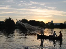 Barco pequeno com pescadores artesanais no Rio Biguaçu ao entardecer, com um deles lançando a rede sobre a água enquanto o sol se põe ao fundo, formando silhuetas. A imagem ilustra a realidade de quem vive da pesca e reforça a importância do Programa Kit Pescador, que oferece equipamentos para mais segurança, proteção e melhores condições de trabalho.
