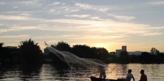 Barco pequeno com pescadores artesanais no Rio Biguaçu ao entardecer, com um deles lançando a rede sobre a água enquanto o sol se põe ao fundo, formando silhuetas. A imagem ilustra a realidade de quem vive da pesca e reforça a importância do Programa Kit Pescador, que oferece equipamentos para mais segurança, proteção e melhores condições de trabalho.