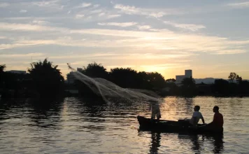 Prefeitura de Biguaçu inicia cadastro para o Programa de Auxílio ao Kit Pescador Barco pequeno com pescadores artesanais no Rio Biguaçu ao entardecer, com um deles lançando a rede sobre a água enquanto o sol se põe ao fundo, formando silhuetas. A imagem ilustra a realidade de quem vive da pesca e reforça a importância do Programa Kit Pescador, que oferece equipamentos para mais segurança, proteção e melhores condições de trabalho.