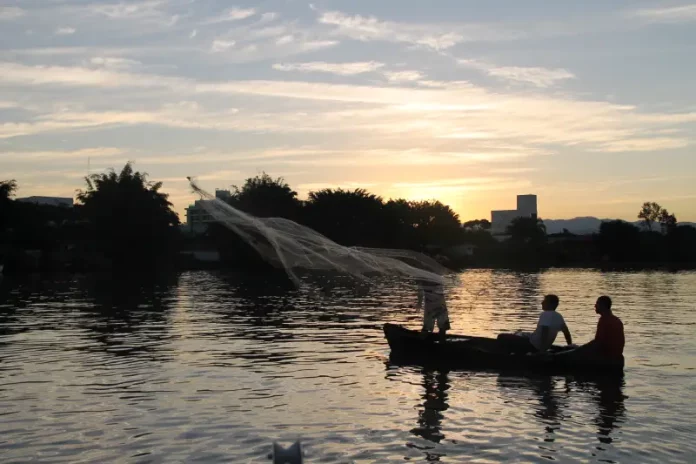 Pescadores-no-Rio-Biguacu Barco pequeno com pescadores artesanais no Rio Biguaçu ao entardecer, com um deles lançando a rede sobre a água enquanto o sol se põe ao fundo, formando silhuetas. A imagem ilustra a realidade de quem vive da pesca e reforça a importância do Programa Kit Pescador, que oferece equipamentos para mais segurança, proteção e melhores condições de trabalho.