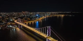 Imagem aérea noturna da Ponte Hercílio Luz iluminada, ligando o continente à Ilha de Santa Catarina, com a finalidade de ilustrar o principal cartão-postal de Florianópolis e destacar sua valorização turística e histórica.