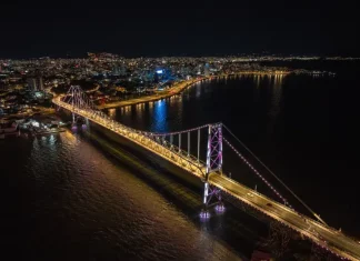 Imagem aérea noturna da Ponte Hercílio Luz iluminada, ligando o continente à Ilha de Santa Catarina, com a finalidade de ilustrar o principal cartão-postal de Florianópolis e destacar sua valorização turística e histórica.