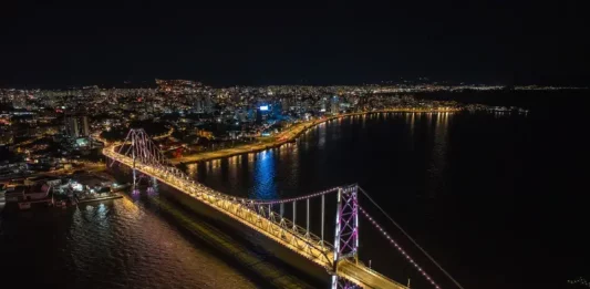Imagem aérea noturna da Ponte Hercílio Luz iluminada, ligando o continente à Ilha de Santa Catarina, com a finalidade de ilustrar o principal cartão-postal de Florianópolis e destacar sua valorização turística e histórica.