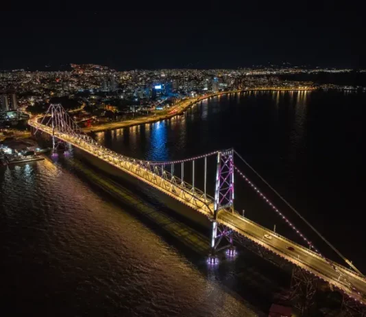 Imagem aérea noturna da Ponte Hercílio Luz iluminada, ligando o continente à Ilha de Santa Catarina, com a finalidade de ilustrar o principal cartão-postal de Florianópolis e destacar sua valorização turística e histórica.
