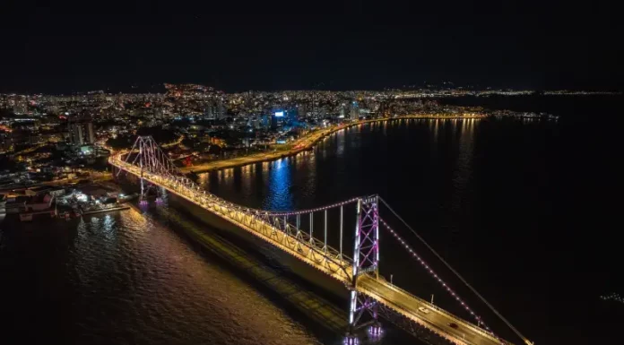 Ponte Hercílio Luz completa 100 anos em 2026 com atrações especiais Imagem aérea noturna da Ponte Hercílio Luz iluminada, ligando o continente à Ilha de Santa Catarina, com a finalidade de ilustrar o principal cartão-postal de Florianópolis e destacar sua valorização turística e histórica.