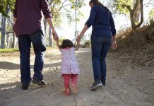 Adultos caminham ao ar livre de mãos dadas com uma criança, simbolizando acolhimento familiar, cuidado, proteção e convivência em ambiente seguro.