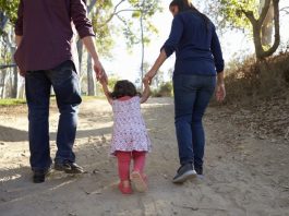 Adultos caminham ao ar livre de mãos dadas com uma criança, simbolizando acolhimento familiar, cuidado, proteção e convivência em ambiente seguro.