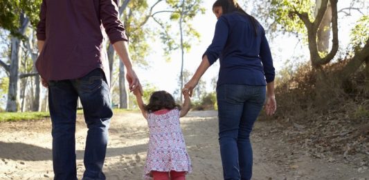 Adultos caminham ao ar livre de mãos dadas com uma criança, simbolizando acolhimento familiar, cuidado, proteção e convivência em ambiente seguro.